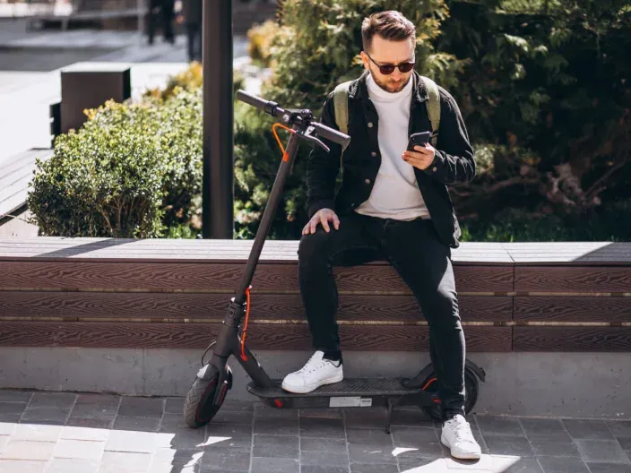 Hombre sentado junto a su patinete eléctrico usando su smartphone en la ciudad, reflejo de la movilidad urbana.