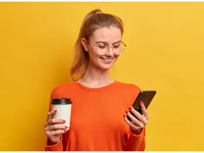 Mujer con gafas usando su móvil y un café sobre fondo amarillo para representar la conectividad y el uso de aplicaciones.