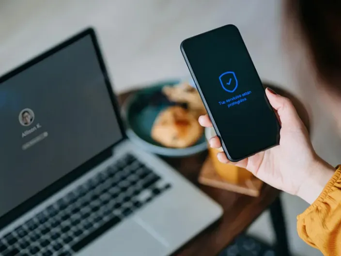 Persona utilizando un ordenador portátil y un teléfono móvil con iconos de seguridad digital en la pantalla.