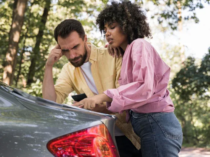 Pareja preocupada junto a un coche averiado en carretera, reflejando la necesidad de asistencia o seguro de auto.