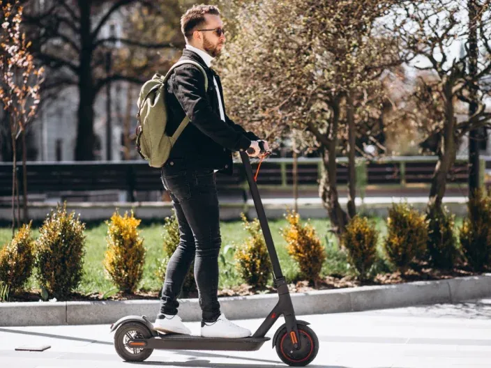 Hombre joven con mochila conduciendo patinete eléctrico negro por un parque como transporte urbano.