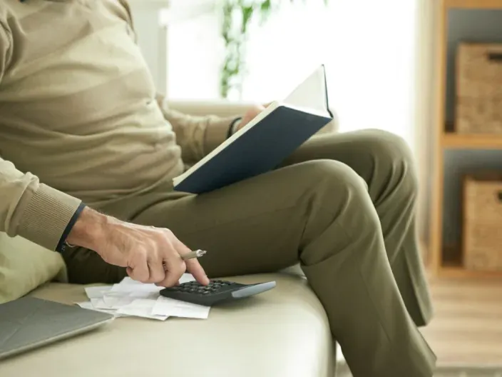 Hombre mayor calculando gastos domésticos con calculadora y libreta para el presupuesto familiar.