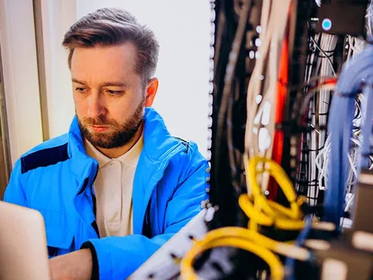 Técnico de telecomunicaciones configurando una red de fibra óptica con un portátil frente a una instalación de cableado estructurado.