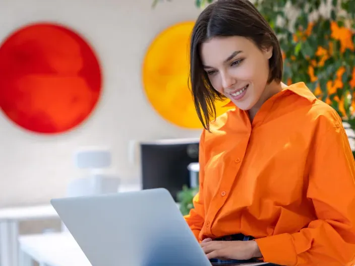 Mujer con camisa naranja usando un portátil en una oficina moderna para representar el teletrabajo y conectividad digital.