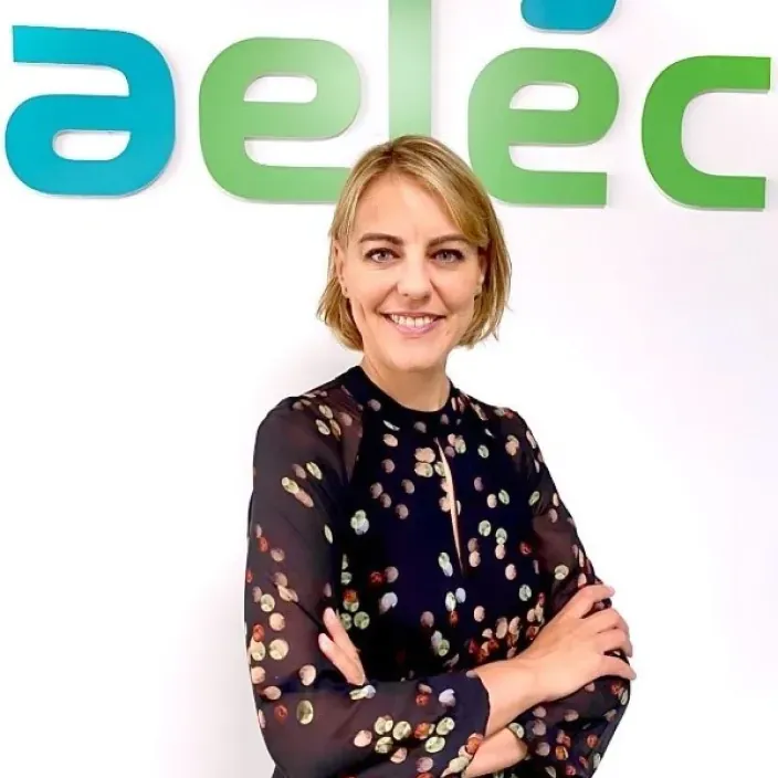 Marta Castro, directora de Regulación de Aelec, posando sonriente con los brazos cruzados frente al logotipo de la asociación eléctrica.