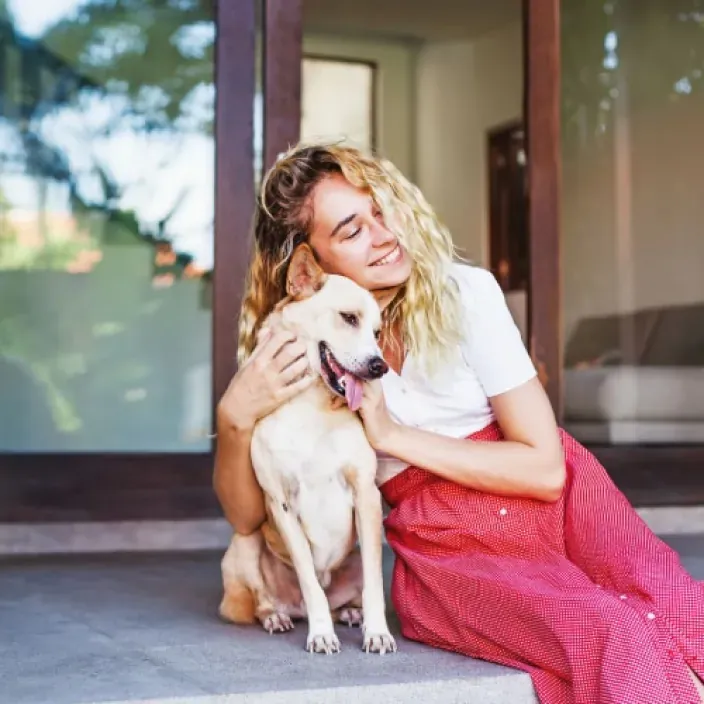 Mujer sonriente abrazando a su perro en la entrada de su casa para representar el bienestar y la protección de mascotas.