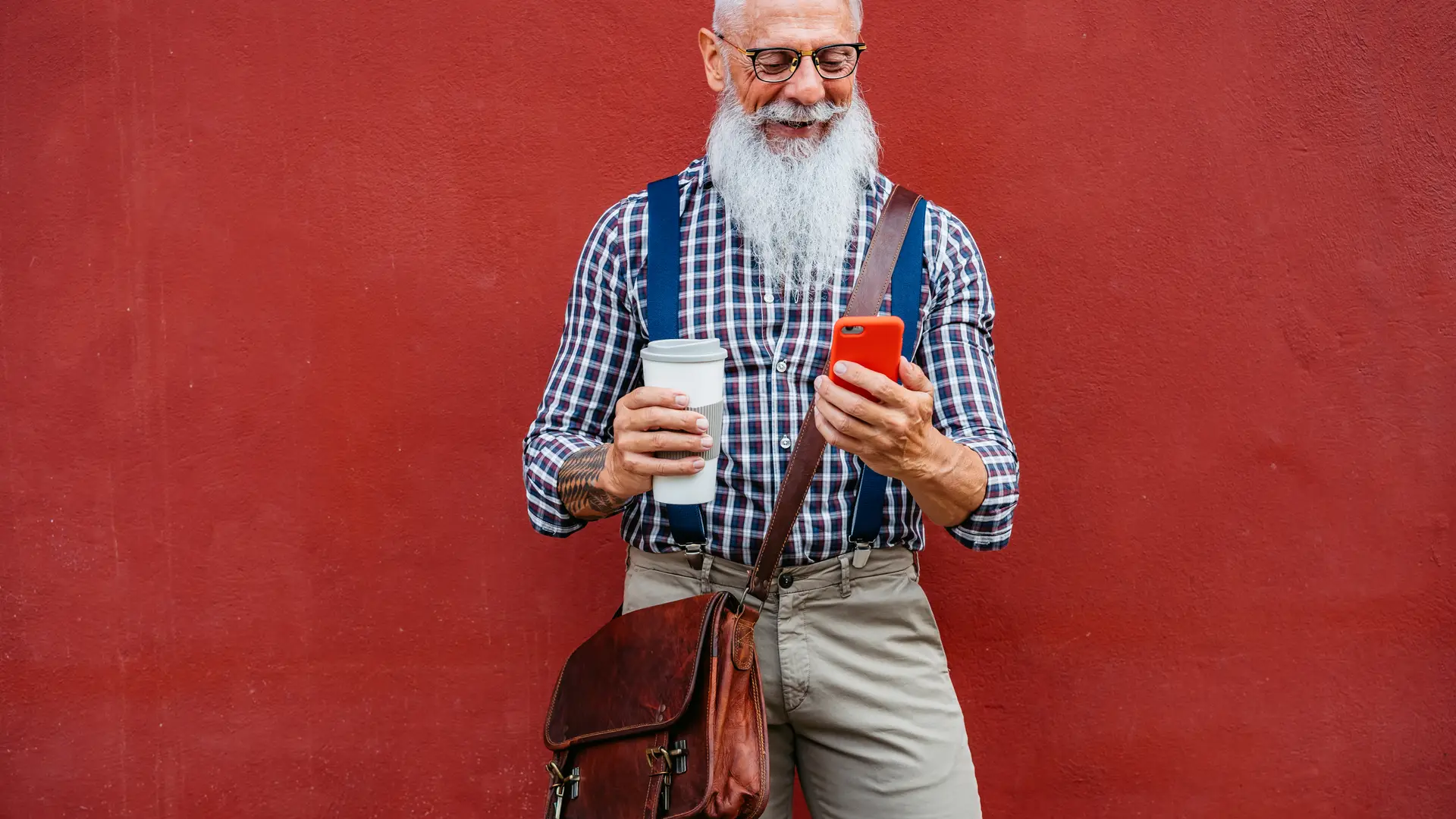 Hombre mayor de estilo hipster consultando su móvil con una pared de fondo de color rojo y un café en la mano en actitud moderna y divertida Hombre mayor de estilo hipster consultando su móvil con una pared de fondo de color rojo y un café en la mano en actitud moderna y divertida