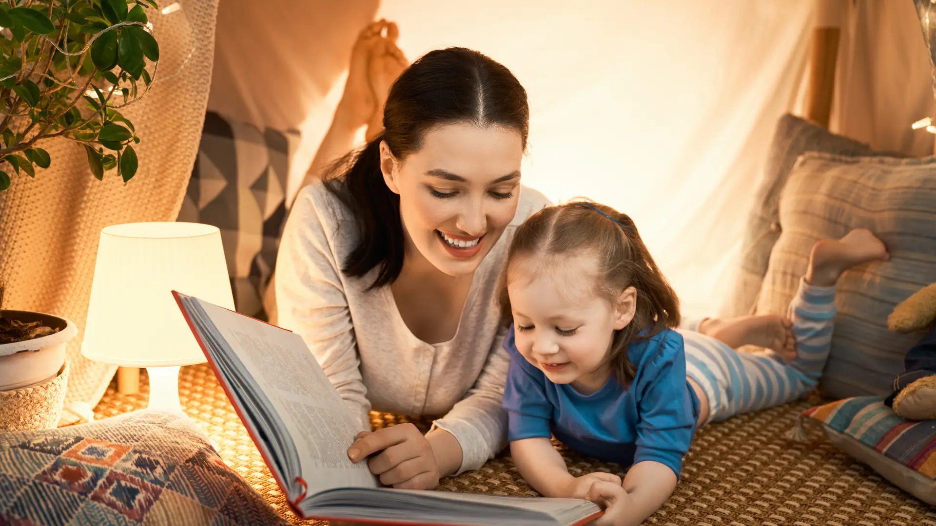 Mujer lee un cuento a su hija pequeña en su refugio construido a base de mantas iluminado gracias a su contrato con Plenitude de la que es cliente Mujer lee un cuento a su hija pequeña en su refugio construido a base de mantas iluminado gracias a su contrato con Plenitude de la que es cliente