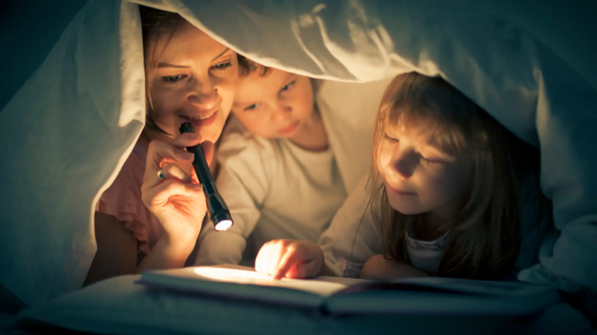 Madre leyendo un libro con sus hijos bajo la manta con la luz de una linterna debido al corte de luz Madre leyendo un libro con sus hijos bajo la manta con la luz de una linterna debido al corte de luz