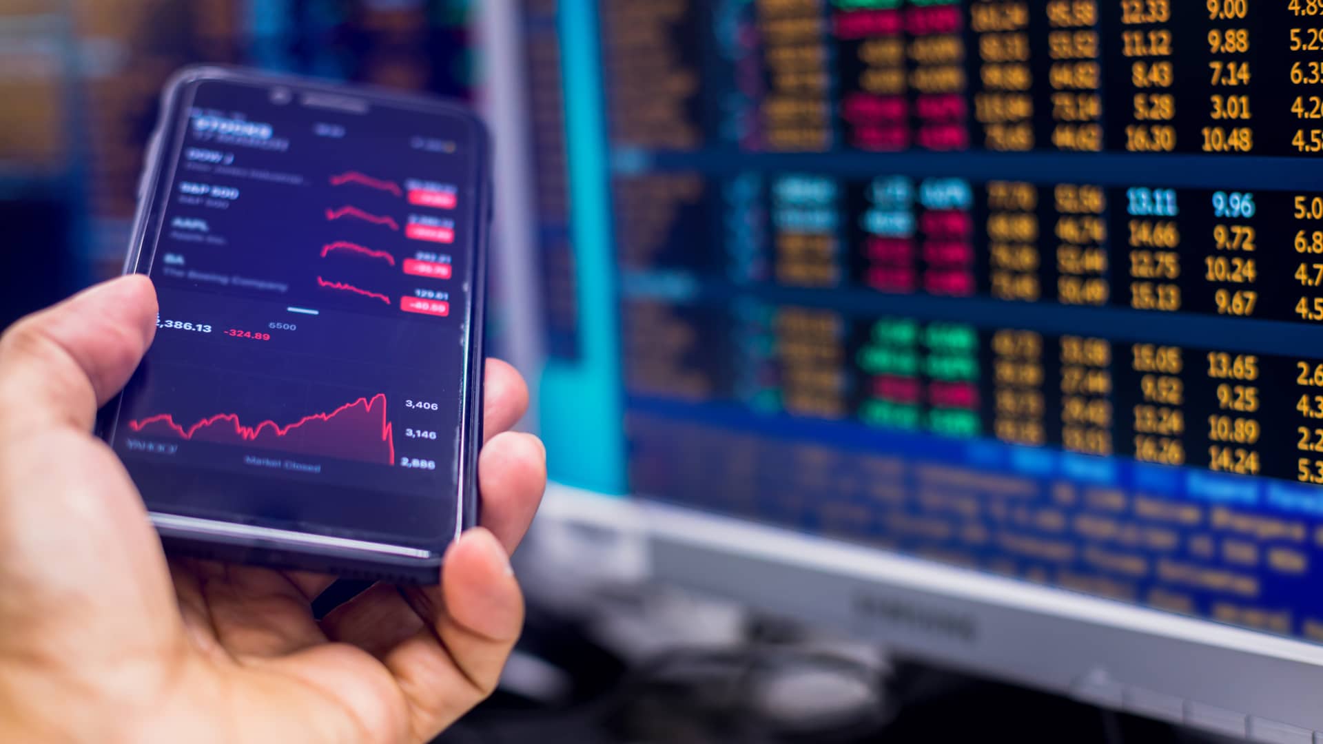 Broker con su smartphone y monitores observando datos bursátiles