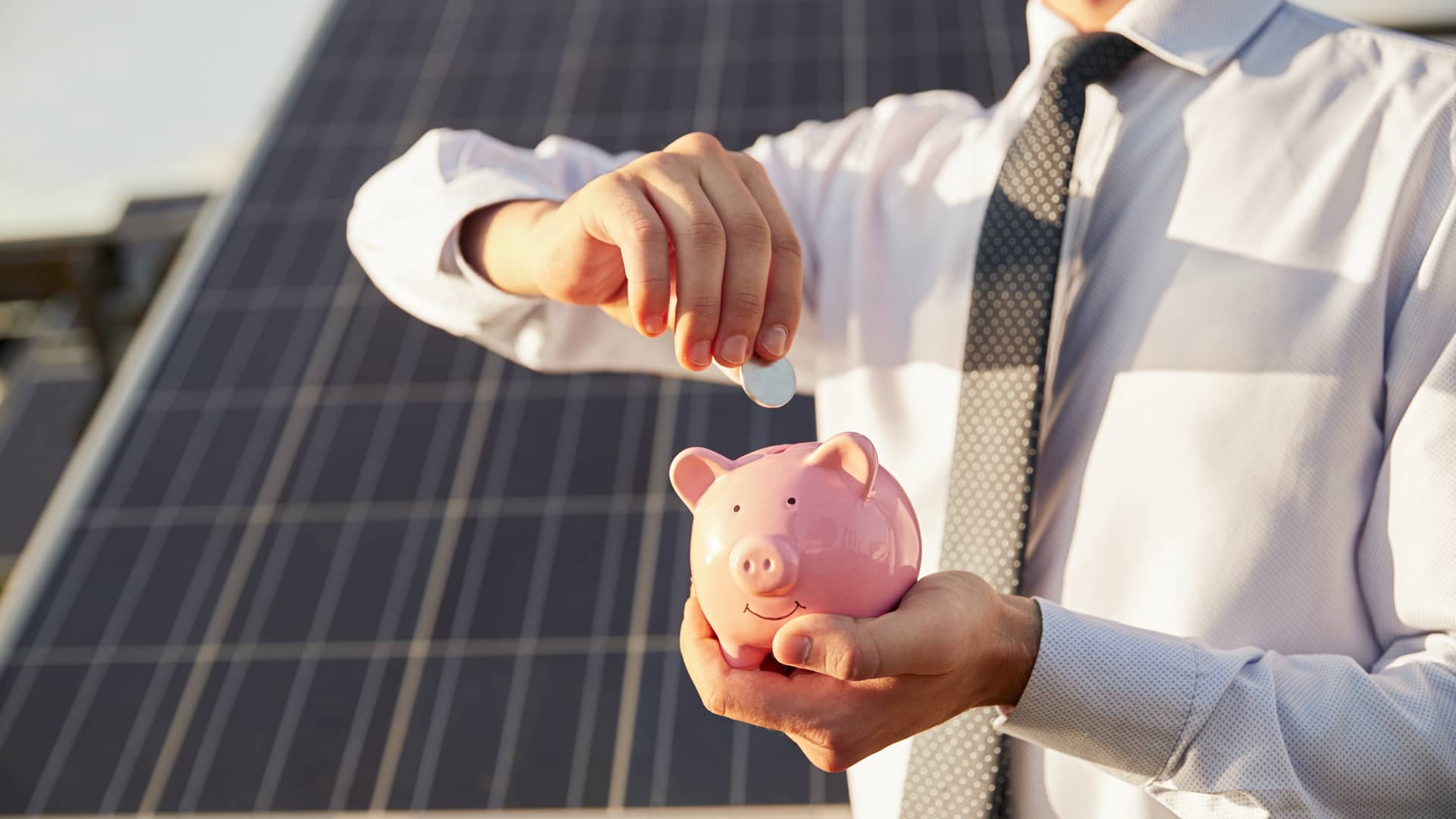hombre metiendo monedas en un cerdito a modo de inversion delante de unas plazas solares que son energias renovables