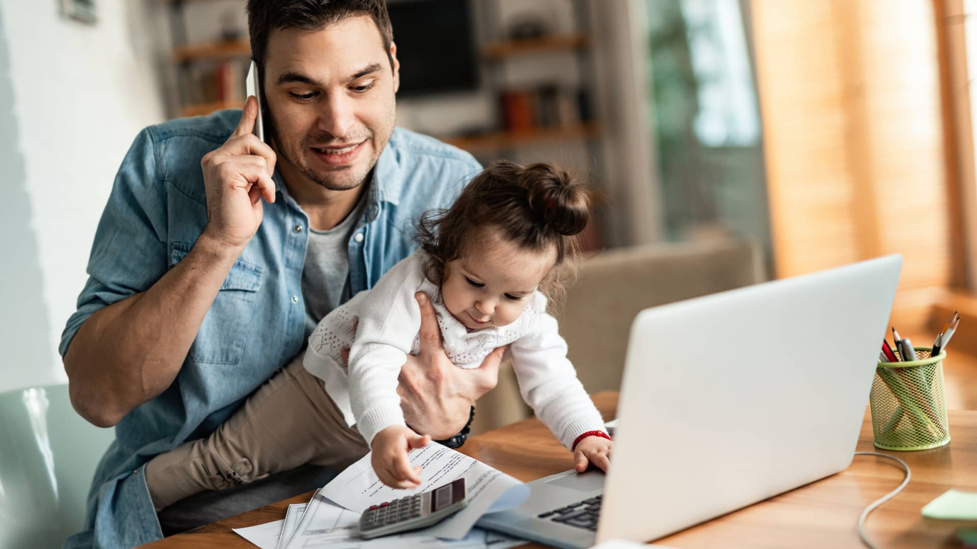 Padre llamando a su seguro Adeslas para solicitar una cita previa para el médico con su hija en brazos jugando con sus papeles Padre llamando a su seguro Adeslas para solicitar una cita previa para el médico con su hija en brazos jugando con sus papeles