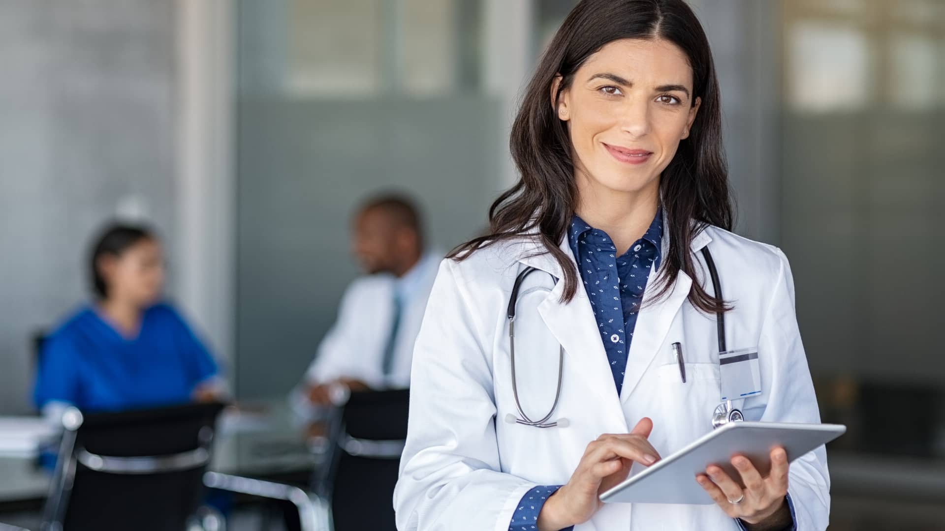 Médico mujer sonriendo a cámara con parte de sus colegas hablando en el fondo como parte del equipo médico del seguro de salud de línea directa