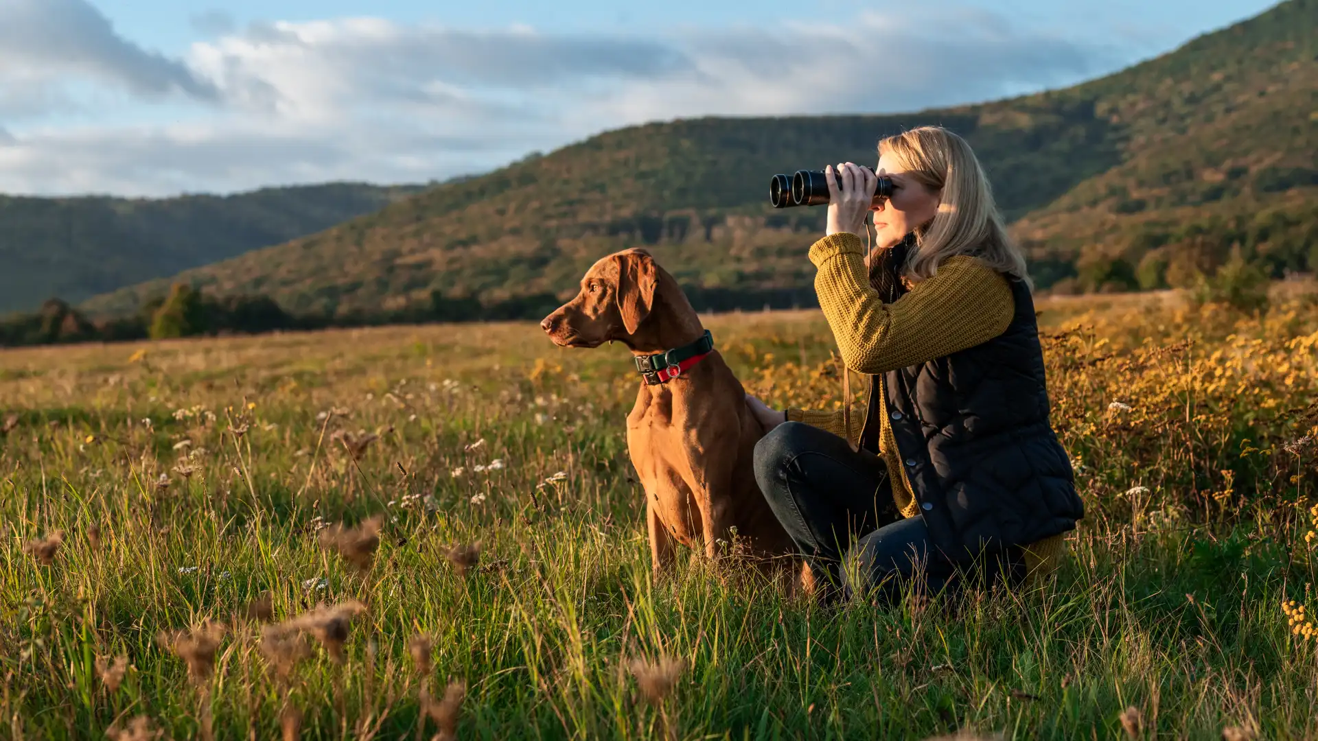 Mujer con prismáticos junto a su perro en una salida de caza, con la tranquilidad de contar con un seguro de caza de mapfre Mujer con prismáticos junto a su perro en una salida de caza, con la tranquilidad de contar con un seguro de caza de mapfre