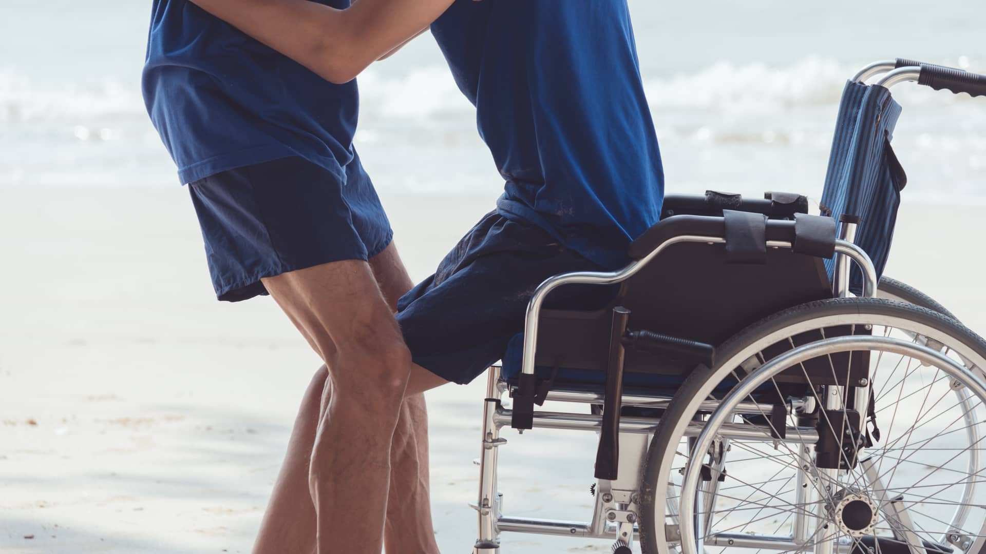 Una persona levantando a un joven de una silla de ruedas que representa el seguro de vida de dependencia de la aseguradora Mapfre Una persona levantando a un joven de una silla de ruedas que representa el seguro de vida de dependencia de la aseguradora Mapfre