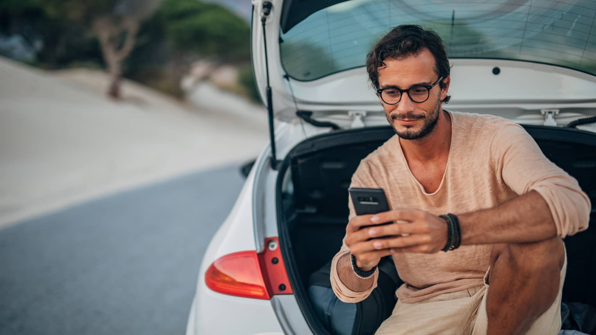 Joven sentando en el maletero de su coche solicitando asistencia en carretera a su seguro con su teléfono móvil