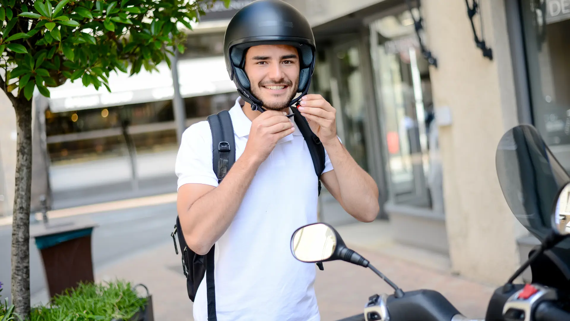Joven utilizando su moto con seguro para repartir pedidos a domicilio, poniéndose el casco Joven utilizando su moto con seguro para repartir pedidos a domicilio, poniéndose el casco