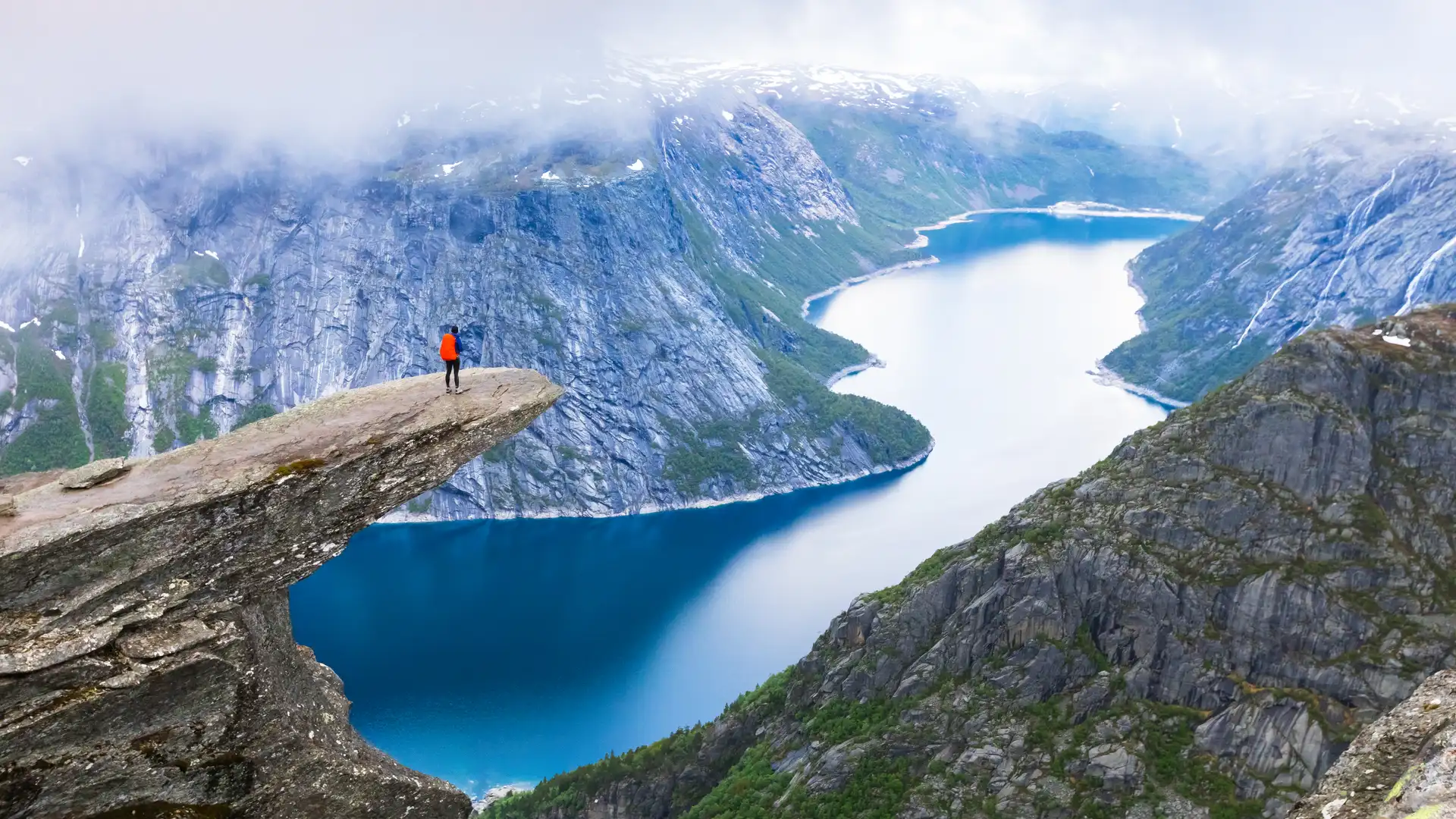 Excursionista de pie en Trolltunga con vista panorámica de lago en Noruega para donde se puede contratar un seguro de viaje Excursionista de pie en Trolltunga con vista panorámica de lago en Noruega para donde se puede contratar un seguro de viaje