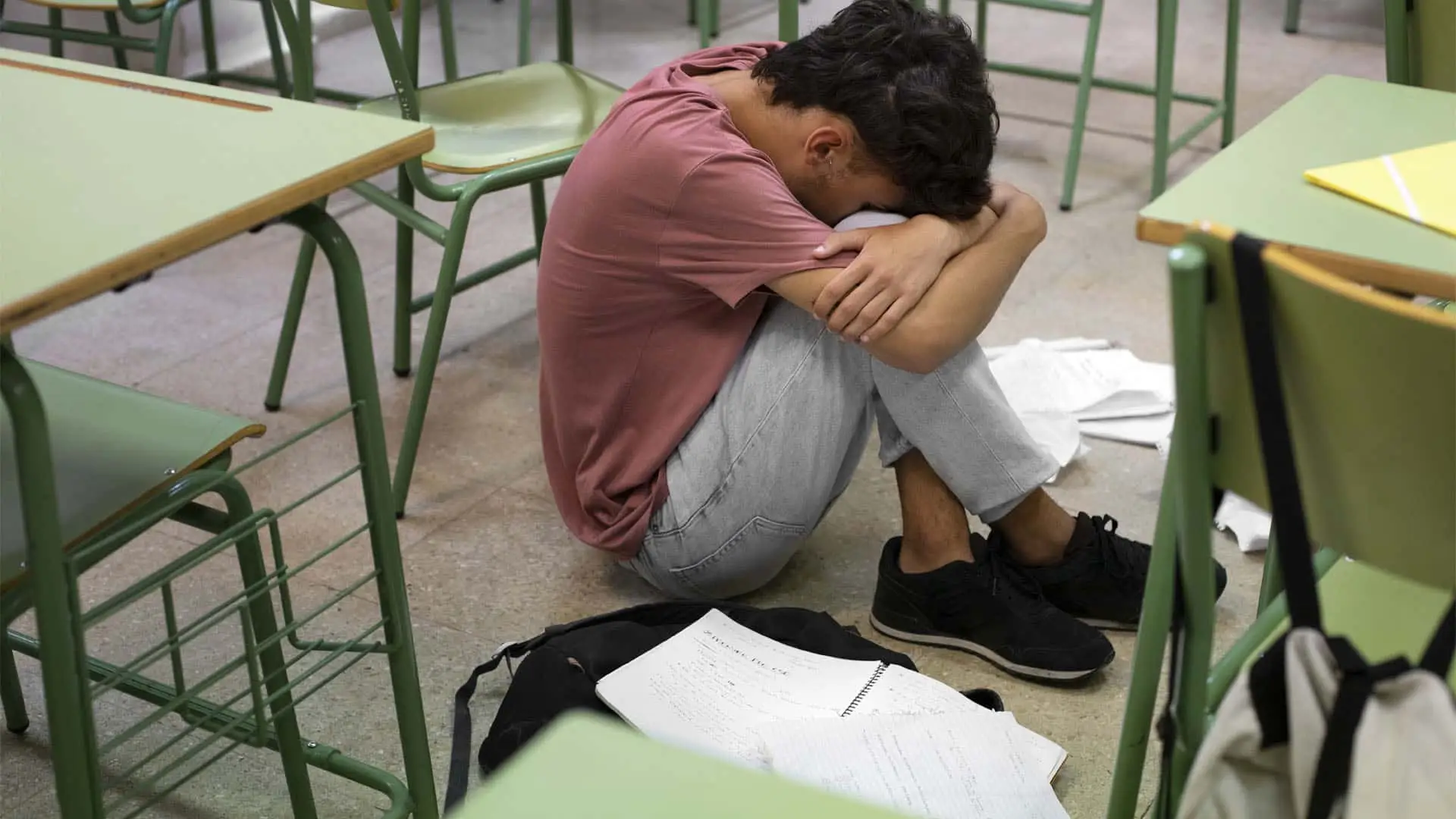 Estudiante sentado en el suelo de un aula, solo y angustiado, como consecuencia del bullying o acoso escolar que sufre.
