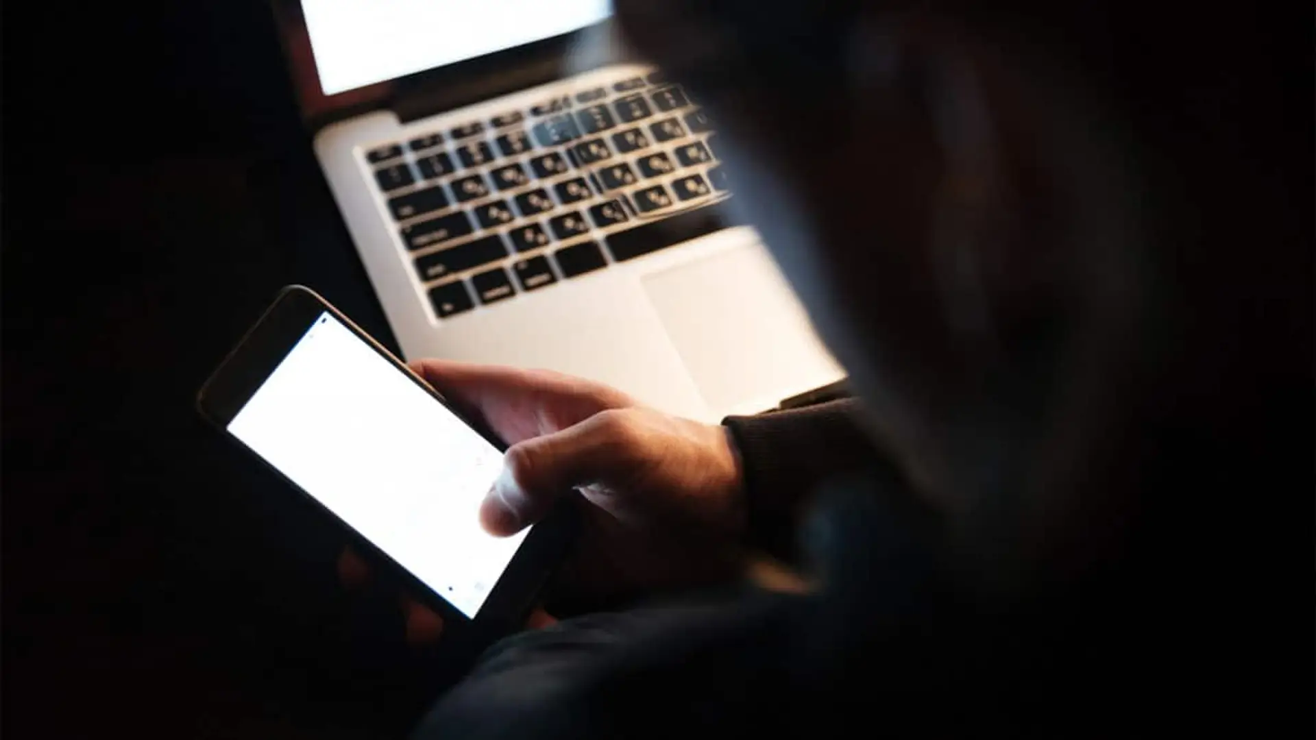 Usuario en un entorno oscuro manipulando su teléfono móvil frente a un ordenador portátil, símbolo de un ciberataque o fraude en línea.