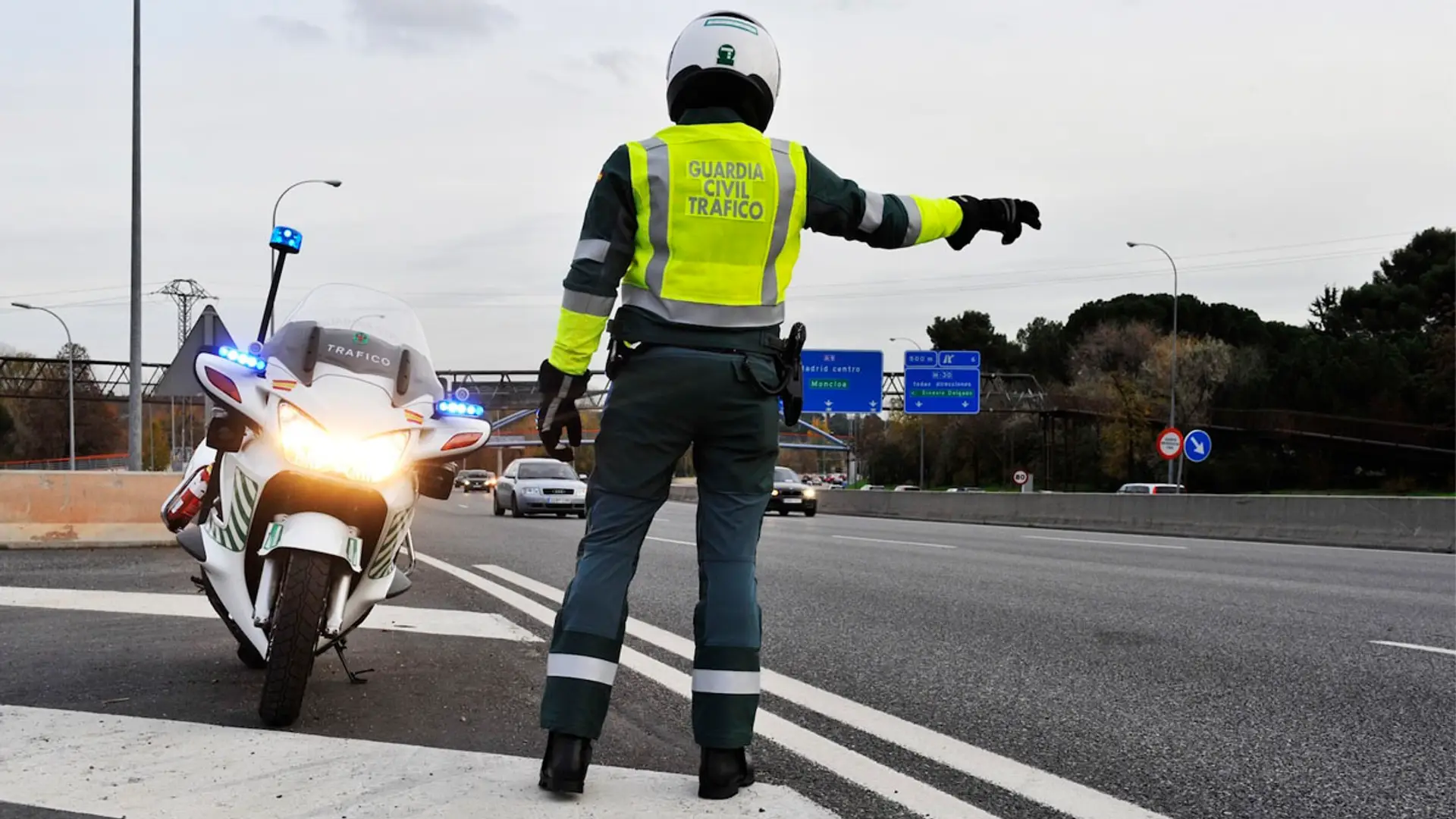 Agente de la Guardia Civil de Tráfico dando el alto a los vehículos en una carretera junto a su motocicleta oficial.