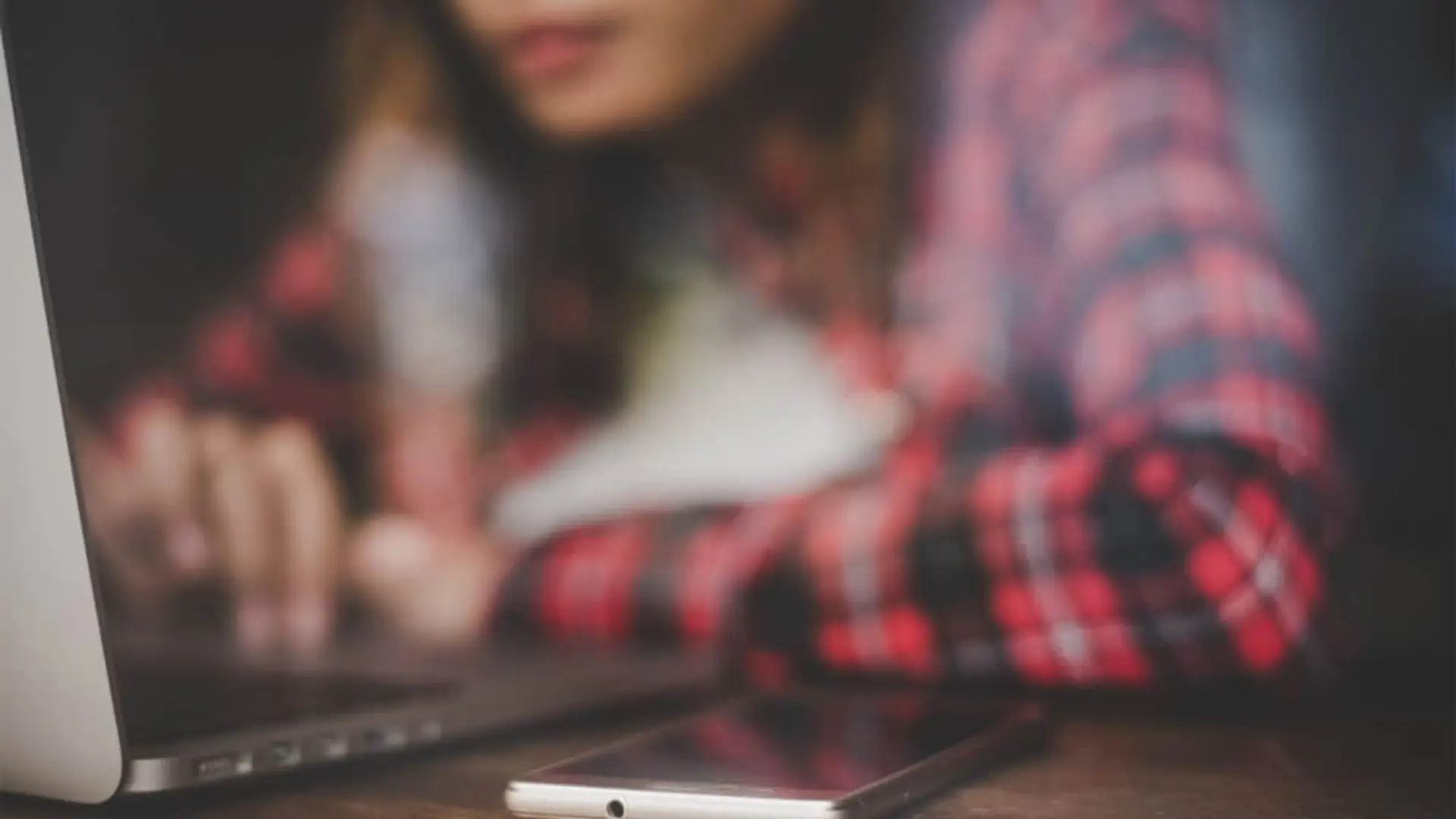 Persona navegando por internet con un ordenador portátil y un teléfono móvil sobre una mesa de madera.