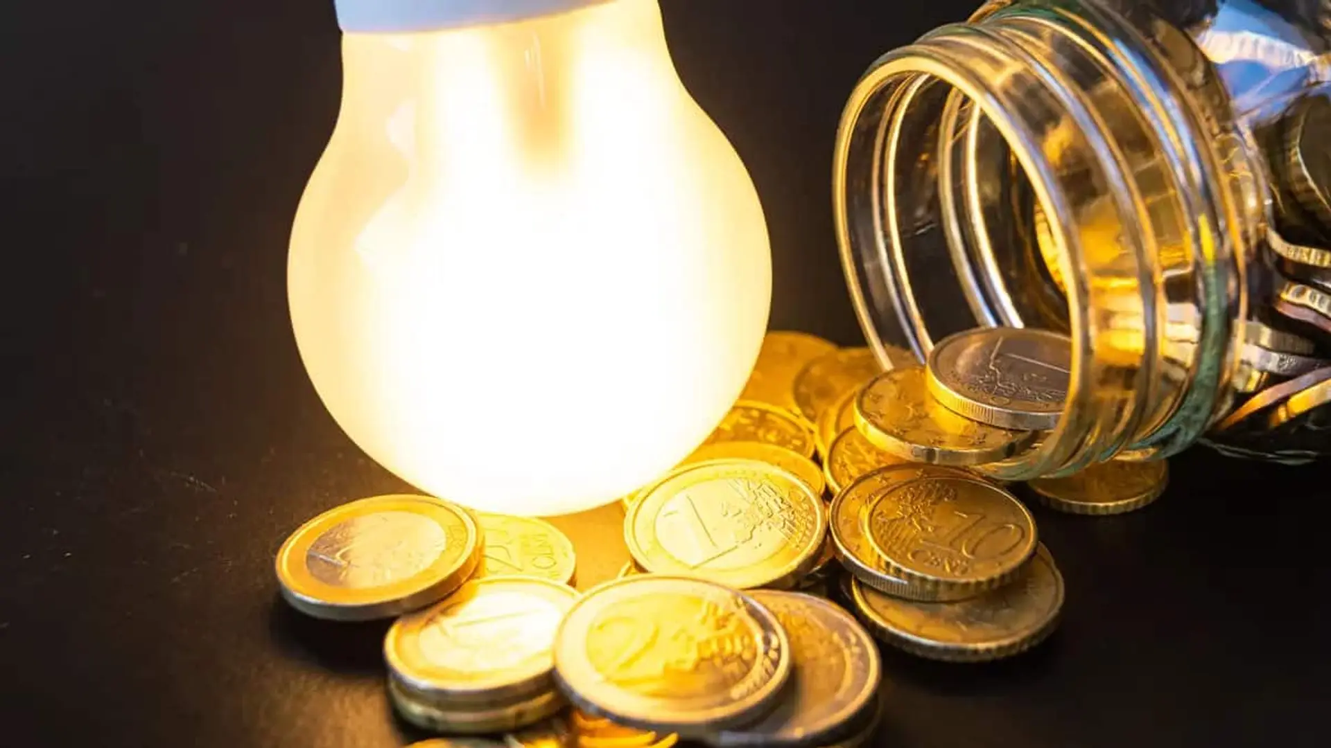 Bombilla encendida junto a un bote de cristal volcado con monedas alrededor.