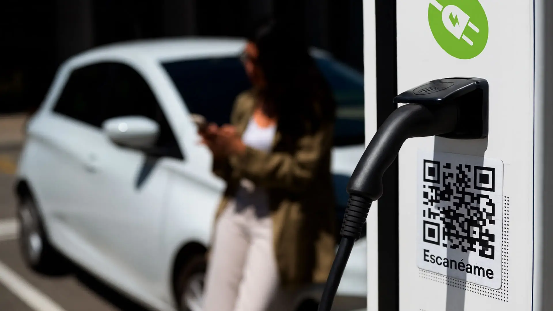 Punto de recarga para coche eléctrico con código QR, con una mujer usando su móvil junto a su vehículo eléctrico.