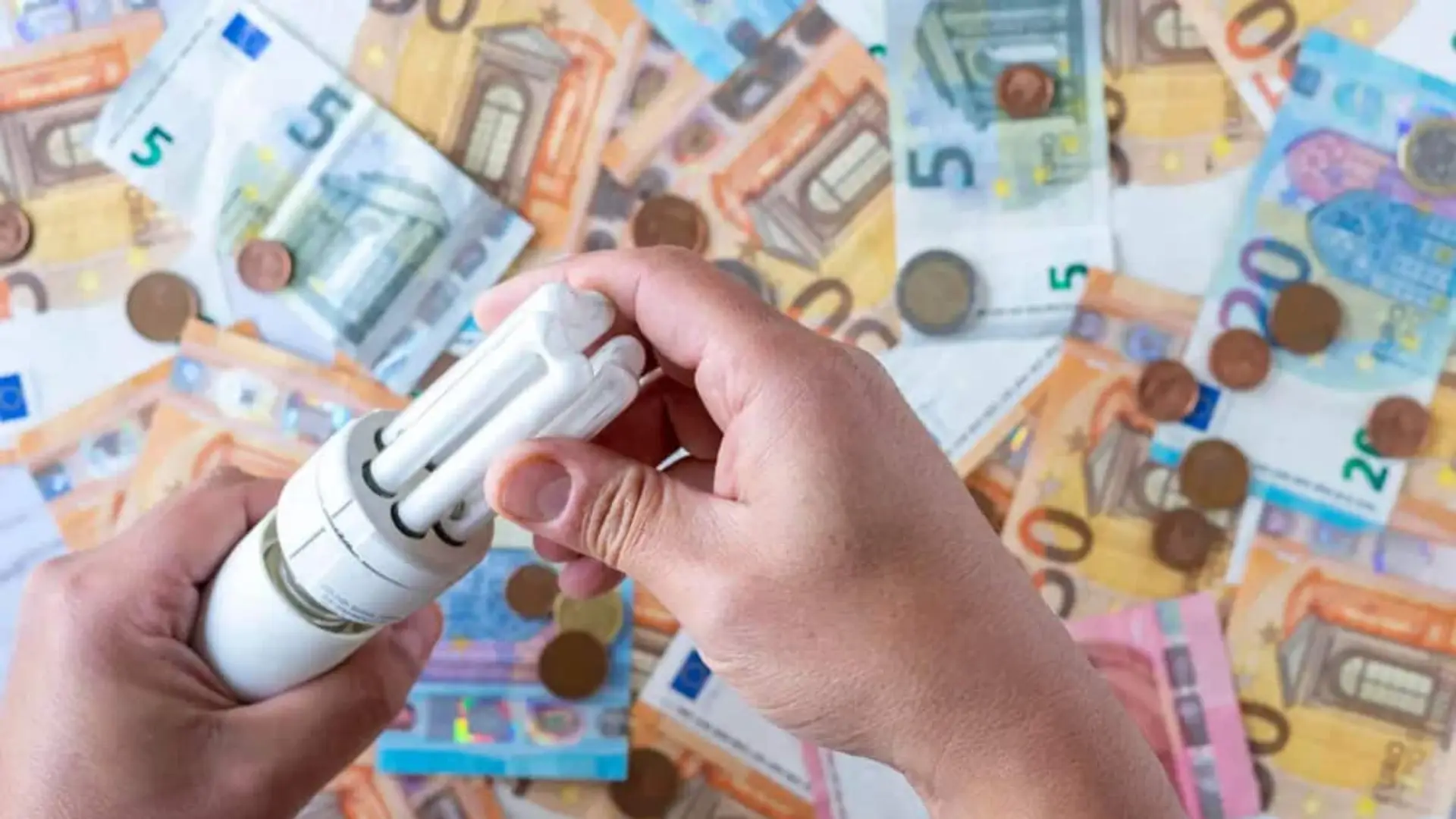10 consejos para reducir (de verdad) tu factura de la luz este otoño Manos sosteniendo una bombilla de bajo consumo sobre una superficie cubierta de billetes y monedas de euro.