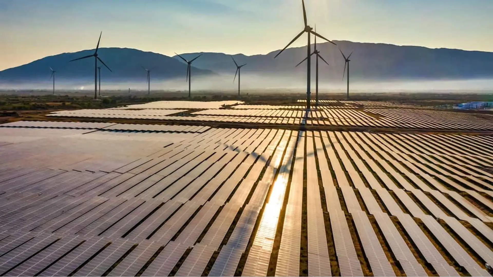 Vista aérea de un parque híbrido con paneles solares y aerogeneradores en una zona montañosa al amanecer.
