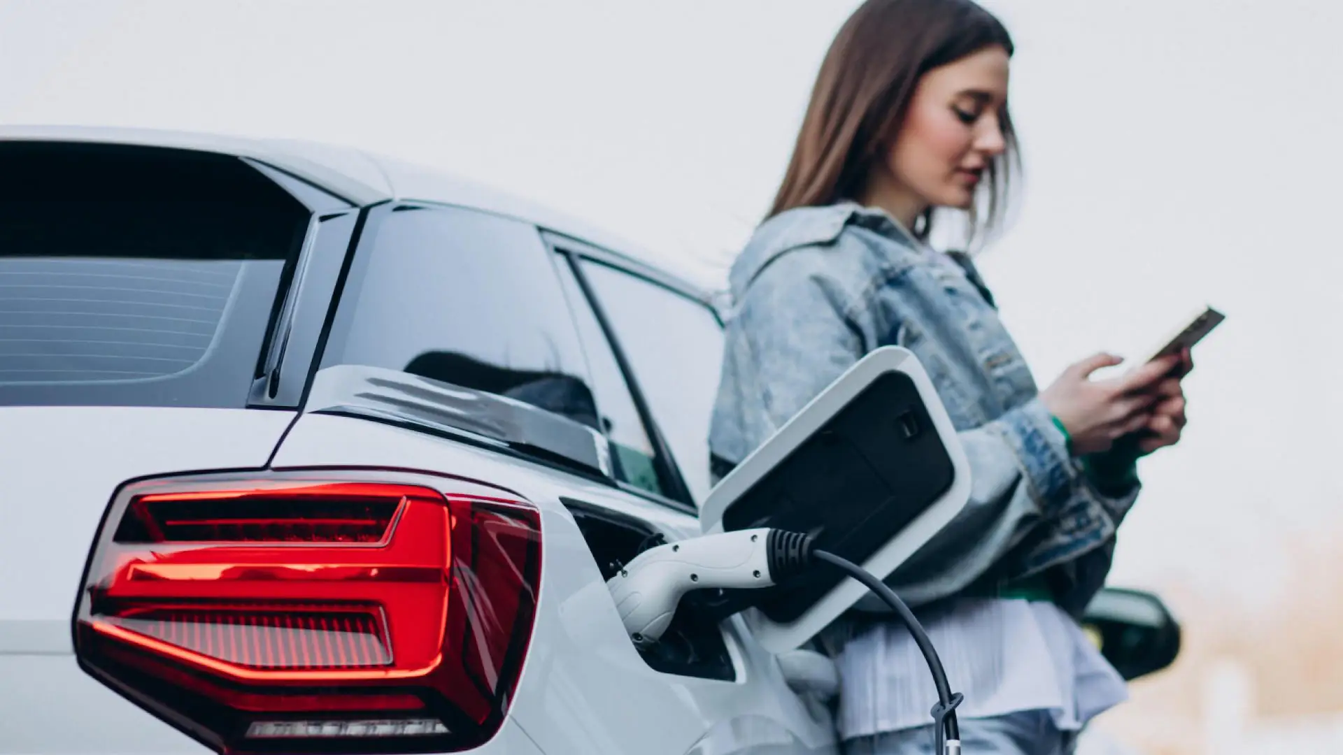 Mujer usando el móvil junto a su coche eléctrico blanco mientras se carga la batería.