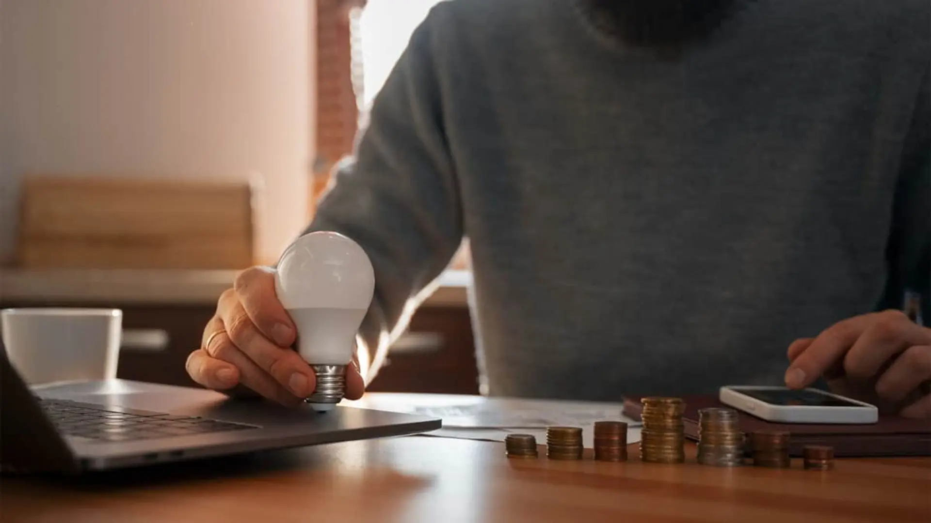 Hombre sosteniendo bombilla LED sobre escritorio con pilas de monedas simbolizando ahorro de luz.