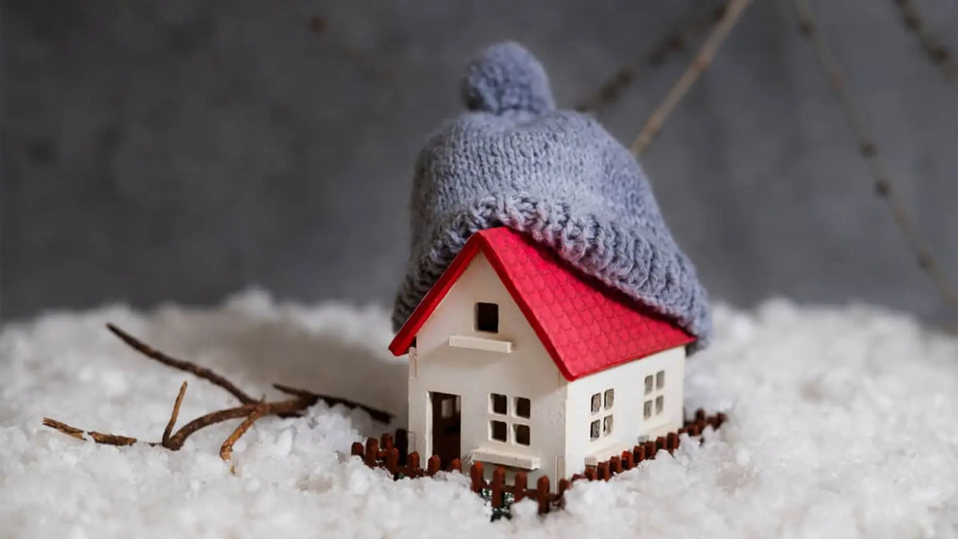 Maqueta de casa cubierta de nieve con gorro de lana gris simbolizando el aislamiento y protección del frío.