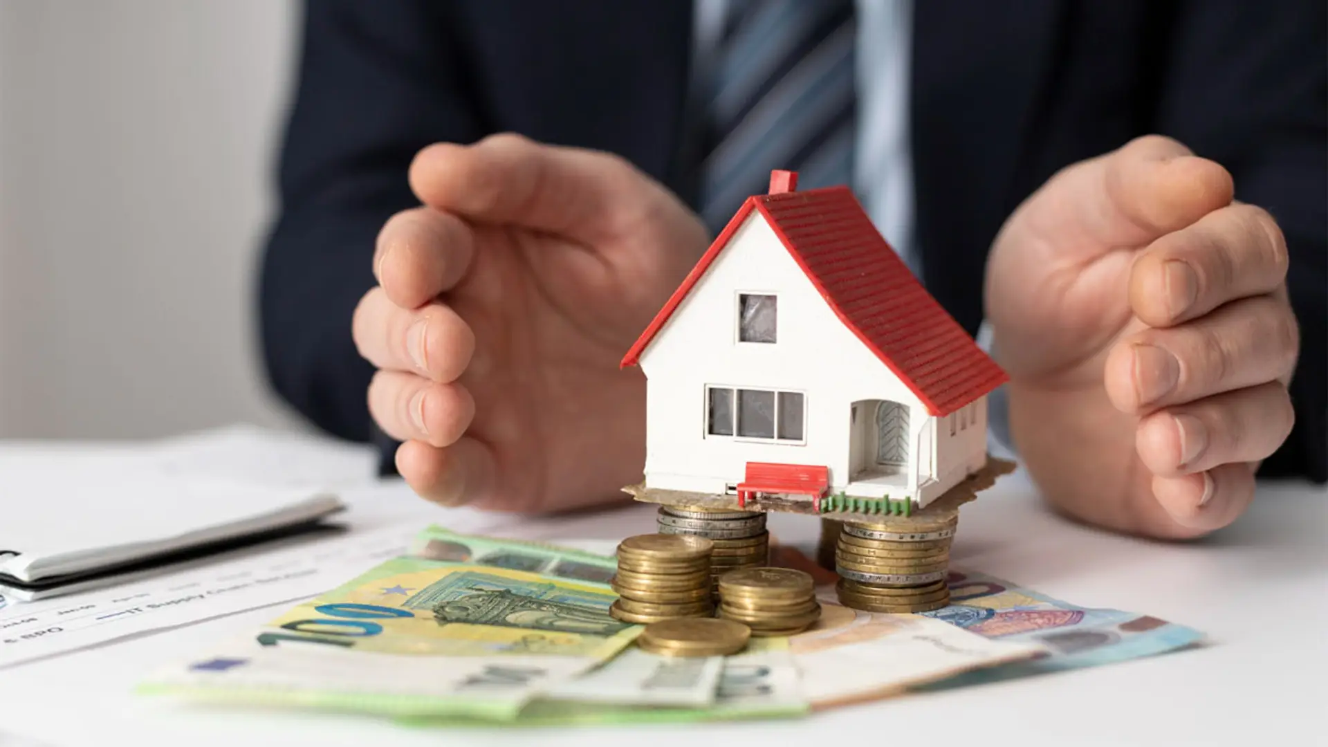 Manos de persona protegiendo una casa sobre monedas y billetes. Concepto de hipoteca.
