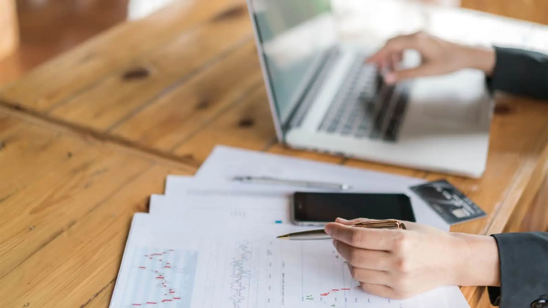 Los depósitos a largo plazo cambian con la llegada de un nuevo banco que paga hasta un 2,8% TAE Persona analizando gráficos financieros en papel con bolígrafo junto a portátil y tarjeta negra.