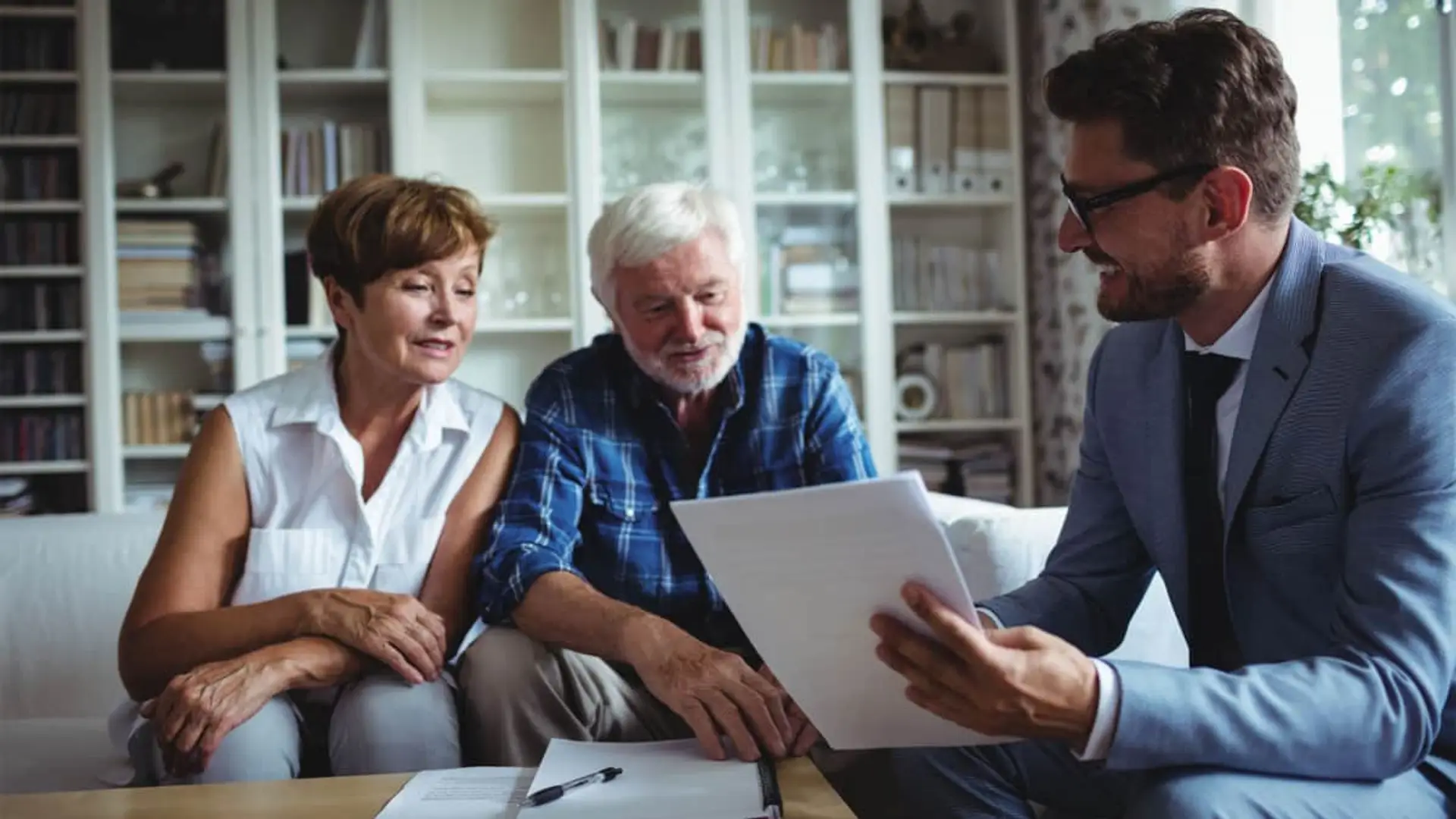 Santander y BBVA reorganizan sus divisiones de seguros y pensiones bajo el “compromiso danés” Asesor financiero explicando documentación de pensión o seguro a pareja de jubilados en casa.