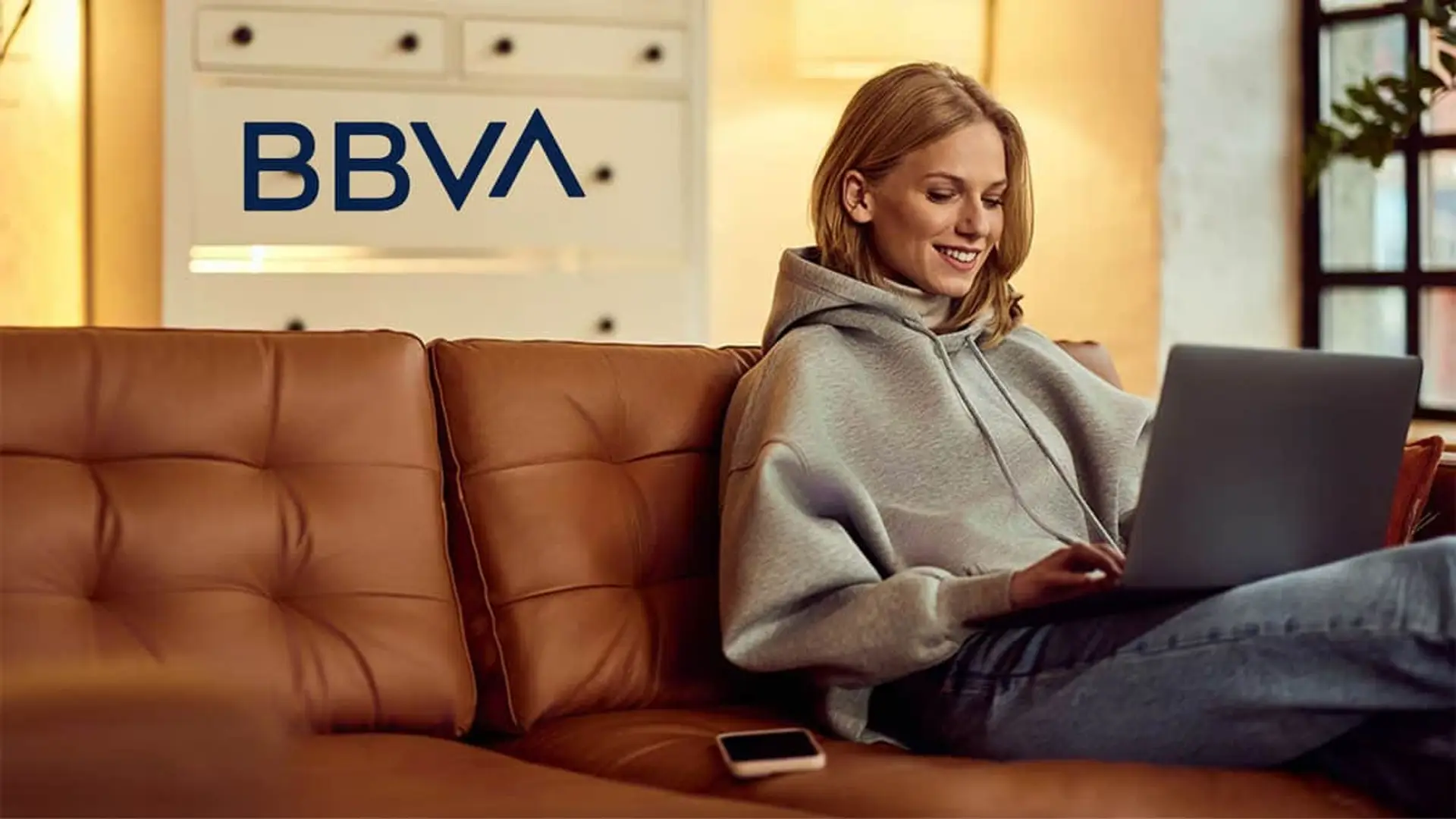 Mujer joven gestionando su banca online de BBVA con un ordenador portátil desde el sofá de su hogar.