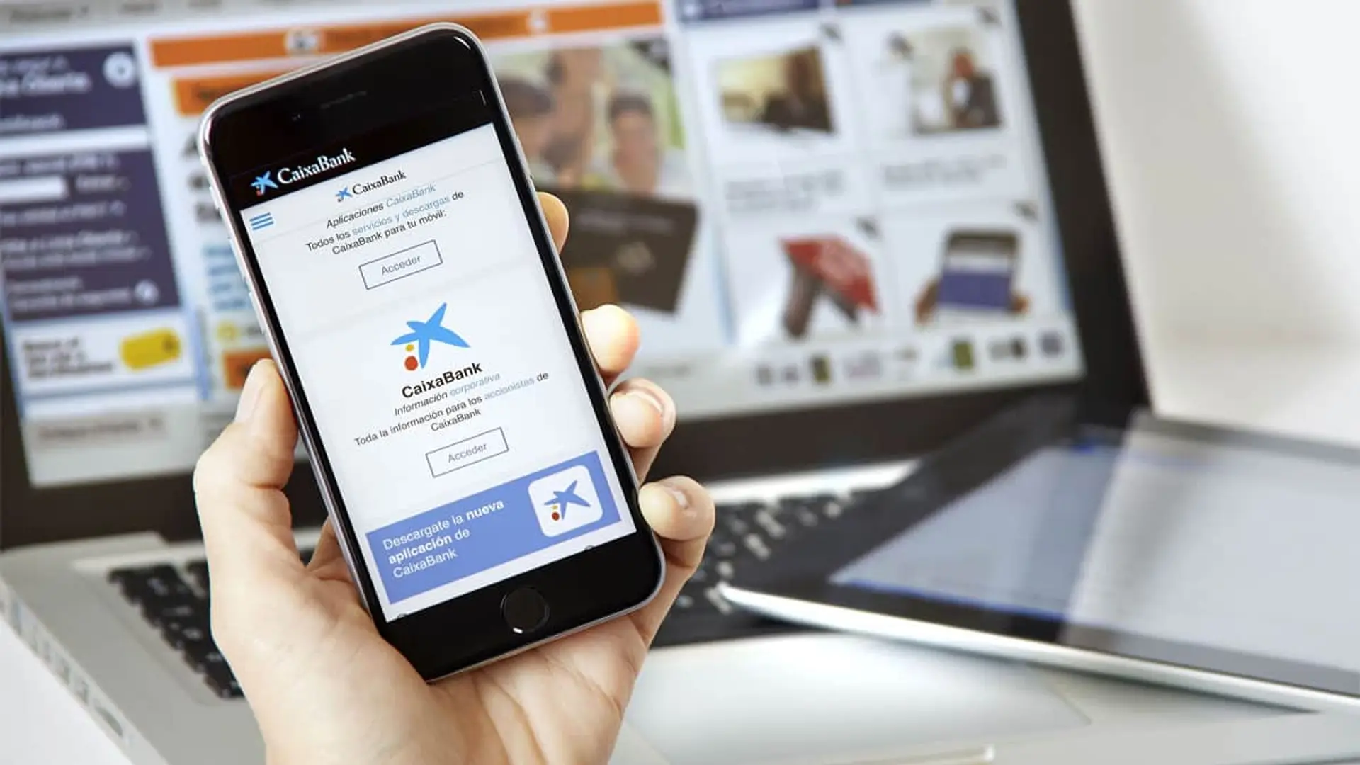 Mano con un smartphone mostrando la aplicación de CaixaBank frente a un ordenador para gestiones de banca digital.