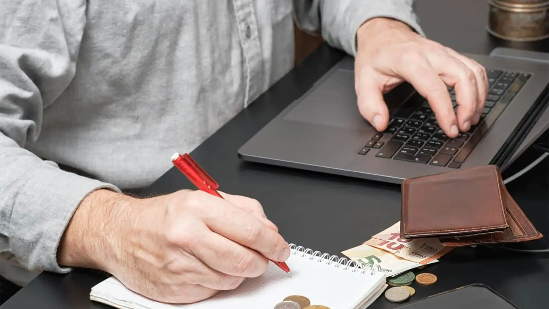 Persona mayor tomando notas en una libreta junto a billetes de euro y un portátil para el control de la economía doméstica.