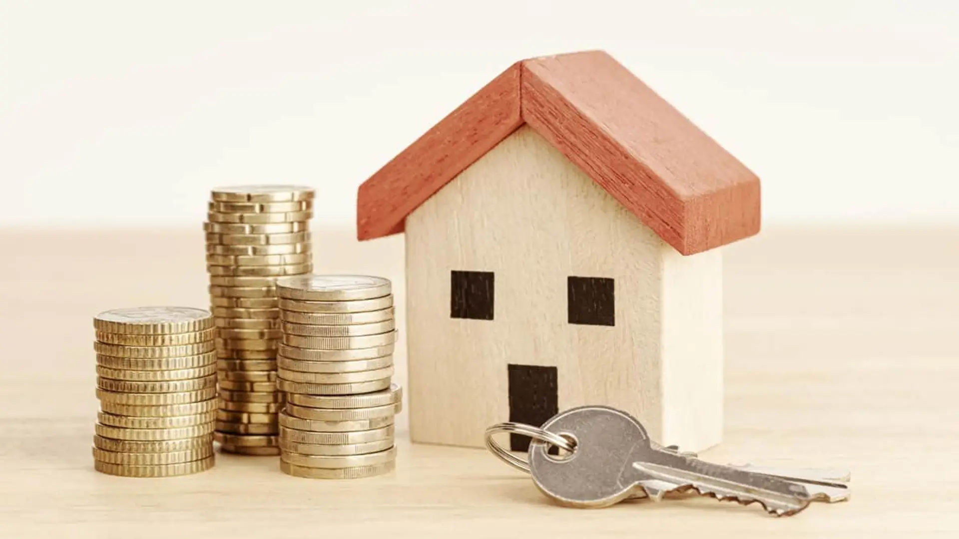 Maqueta de casa de madera junto a pilas de monedas y llaves simbolizando la hipoteca o compra.