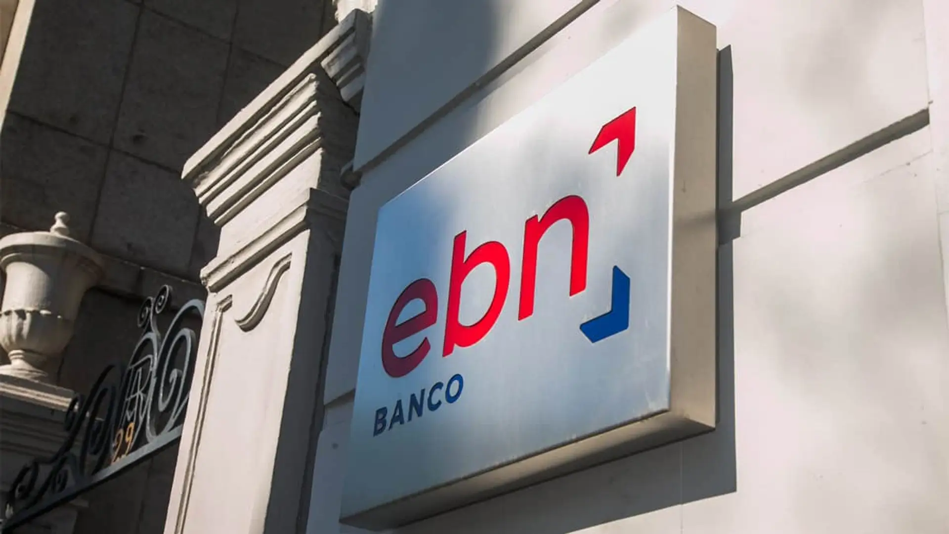 Rótulo luminoso con logotipo de EBN Banco en la fachada de piedra de una sucursal bancaria.