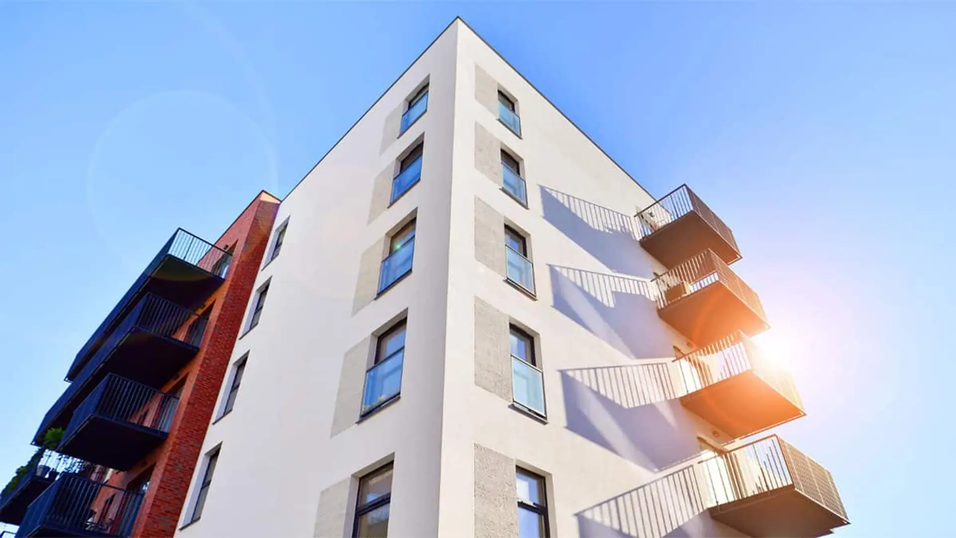 Edificio de apartamentos moderno con balcones visto desde abajo y cielo despejado de fondo.