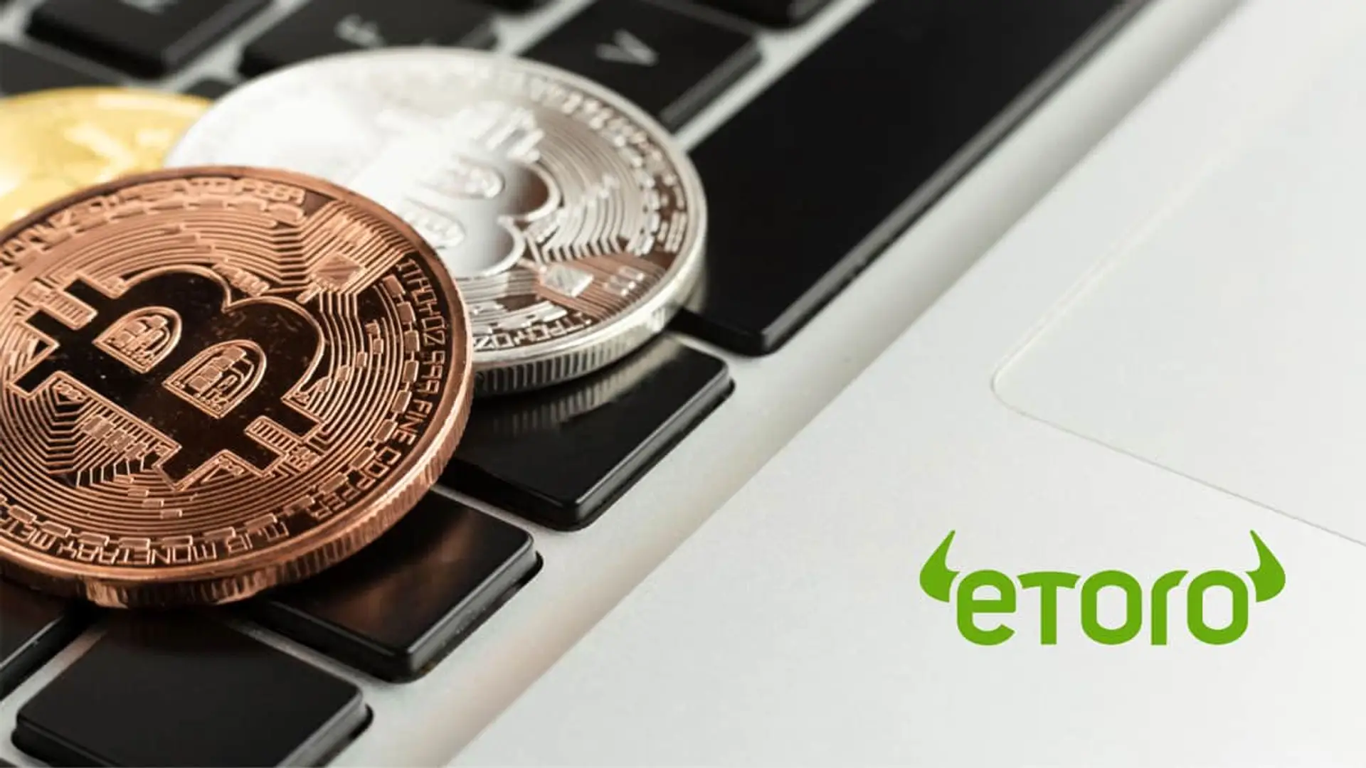 Monedas físicas de Bitcoin sobre teclado de portátil junto al logo de la plataforma de trading eToro.