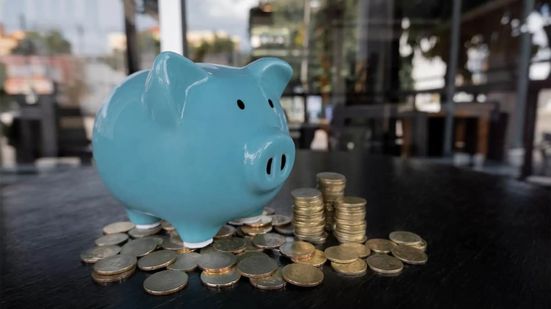 Hucha cerdito azul rodeada de pilas de monedas sobre mesa de madera simbolizando el ahorro.