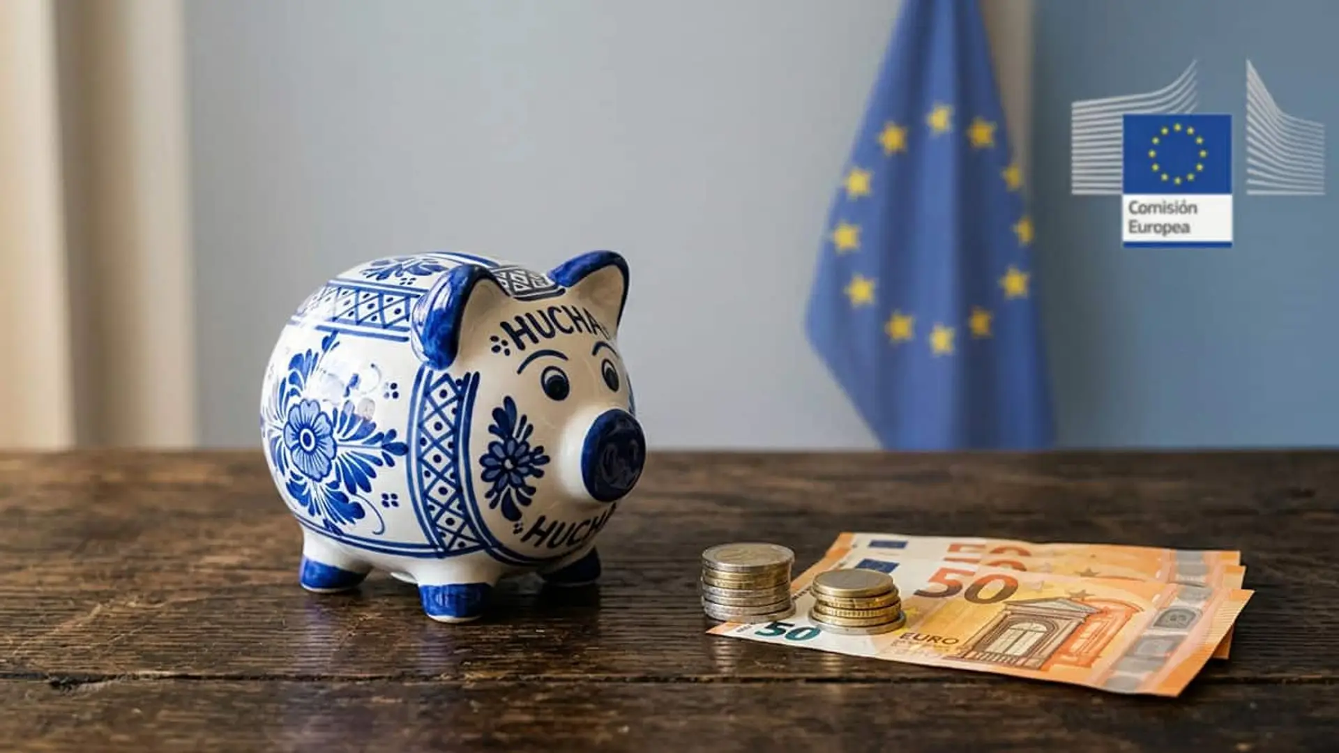 Hucha con forma de cerdito junto a monedas y billetes de euro con la bandera de la Unión Europea y la Comisión Europea al fondo