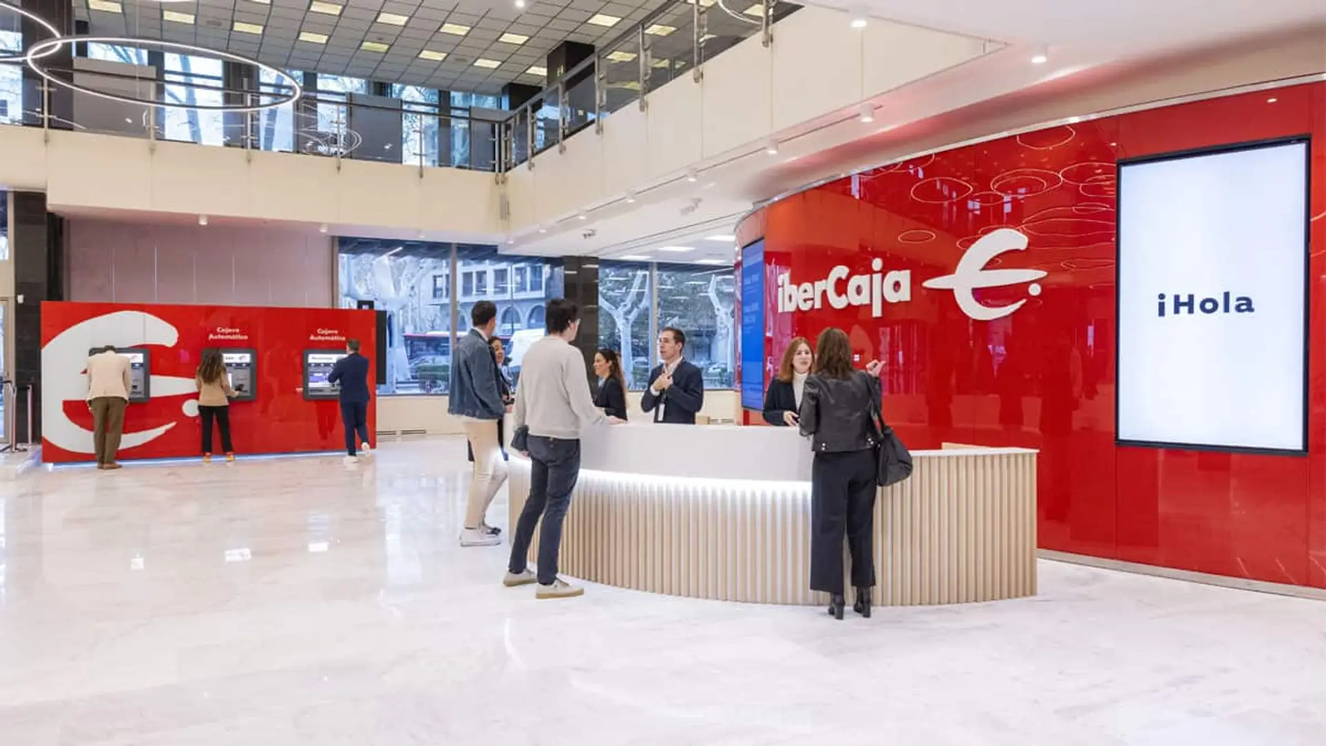 Zona de atención al cliente en una oficina moderna de Ibercaja con mostradores, empleados y cajeros automáticos al fondo.