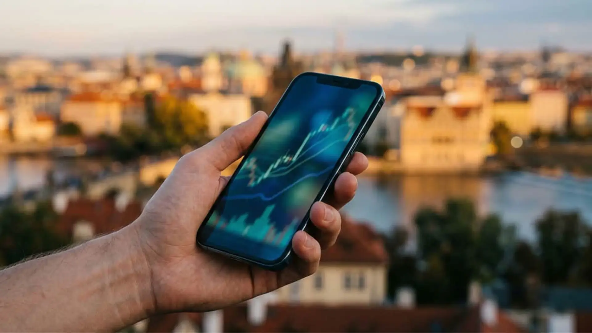 Mano sosteniendo un móvil con gráficos bursátiles de velas japonesas frente a un paisaje urbano europeo al atardecer