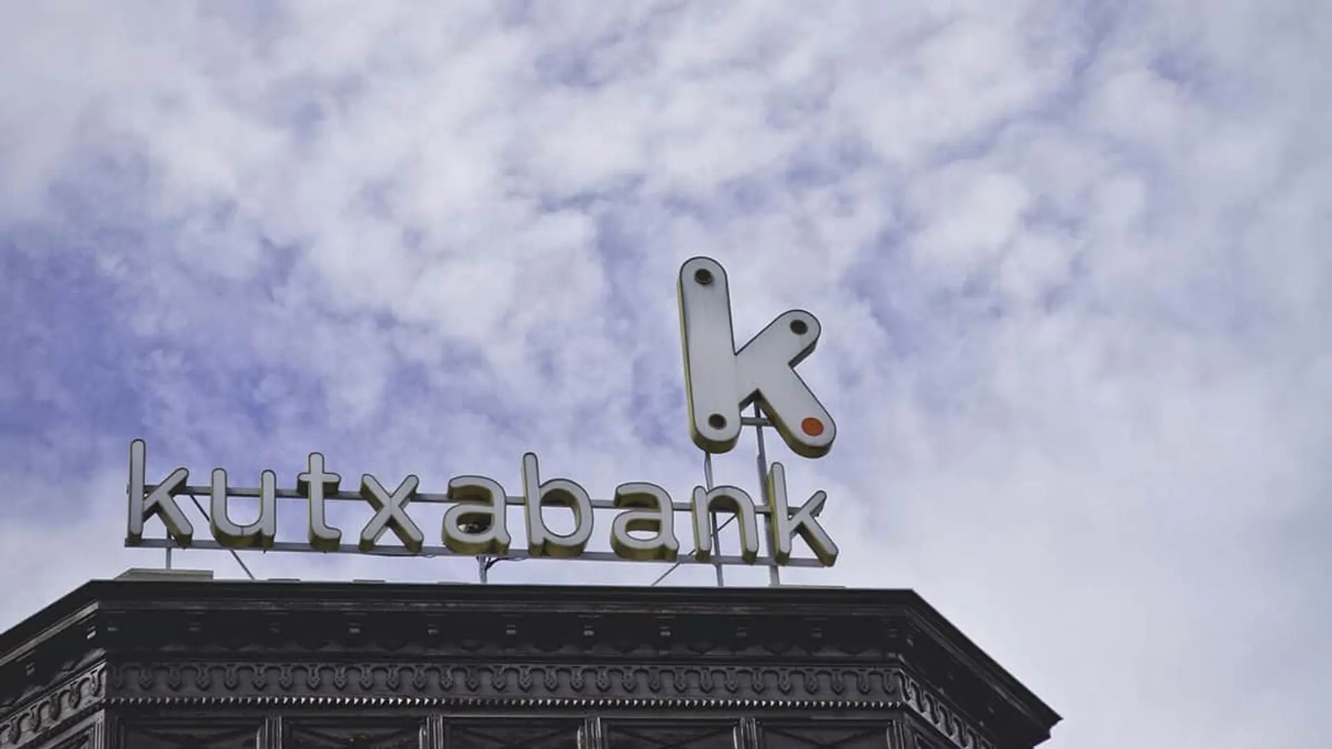 Logotipo de Kutxabank en lo alto de un edificio corporativo sobre un fondo de cielo nublado.