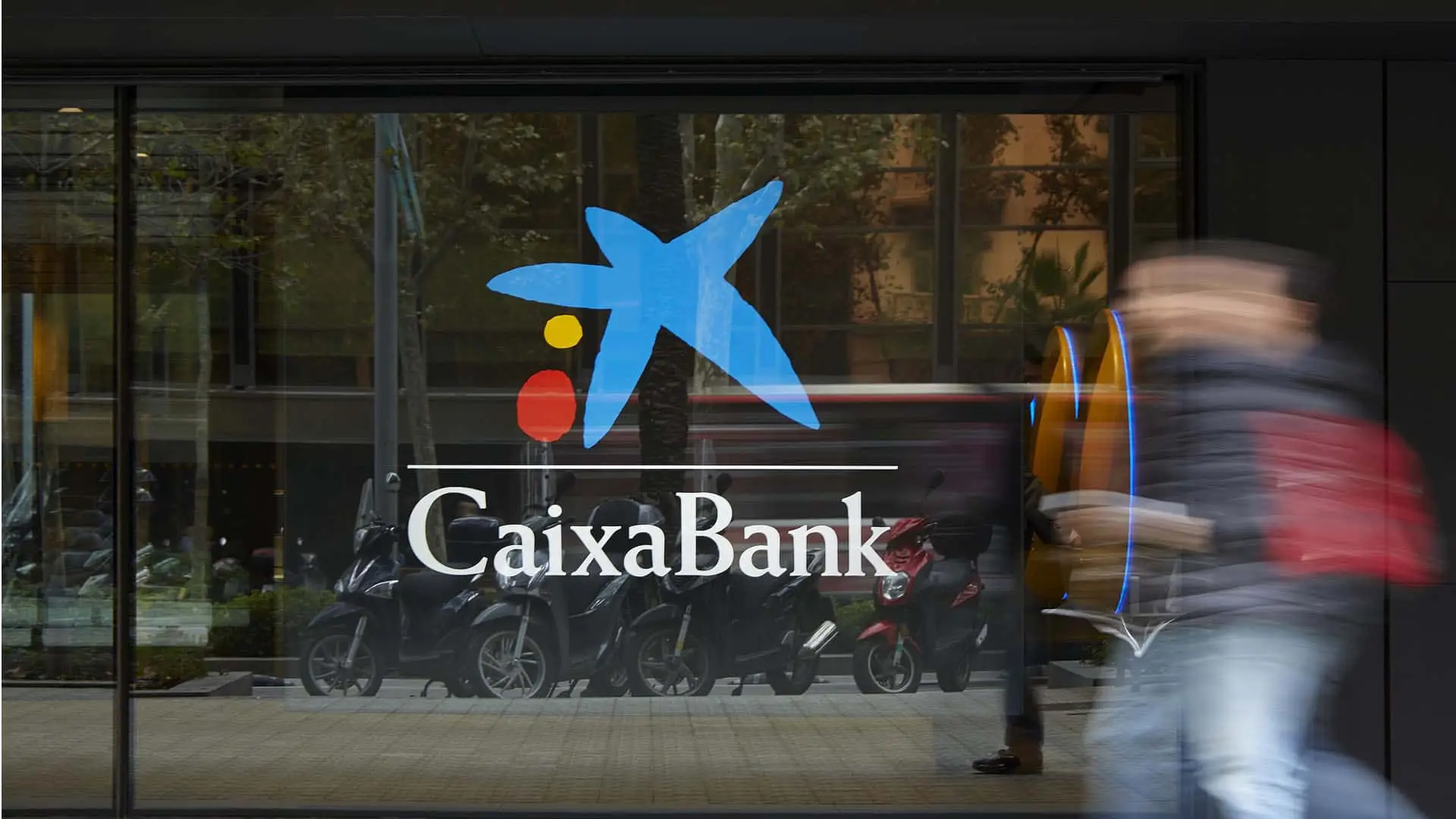 Escaparate con el logotipo de CaixaBank en un cristal con personas caminando frente a la oficina.