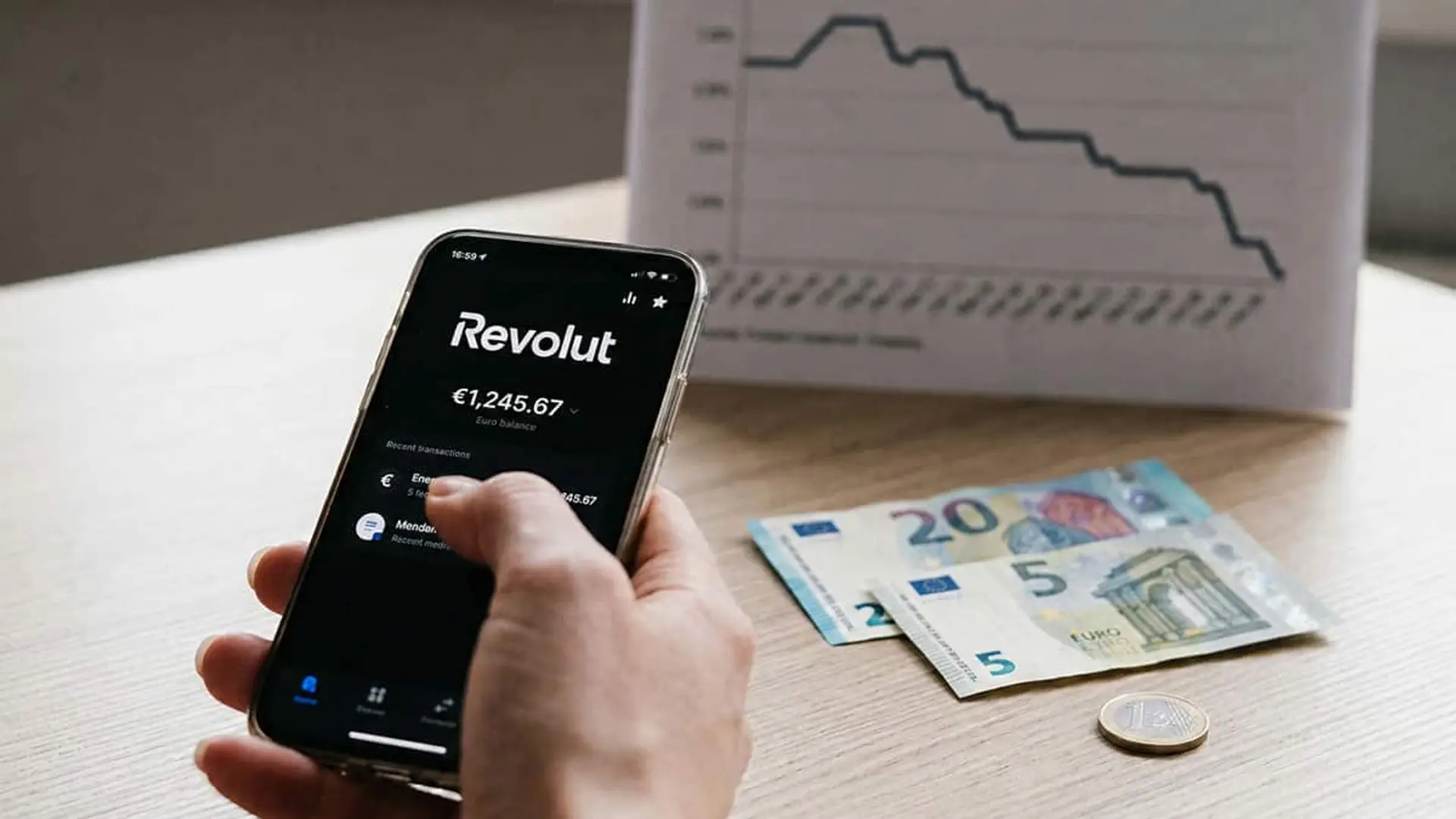 Mano sosteniendo un móvil con la aplicación de Revolut junto a billetes y monedas de euro y un gráfico descendente